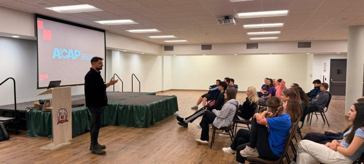 (English) A guest speaker stands beside a podium and presents to Klub Stepinac teens seated on chairs in the parish hall, with the ACAP (Association of Croatian American Professionals) slide on the screen behind him.