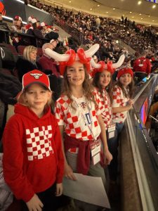 Four girls in red-and-white checkered šahovnica shirts and Bulls hats, two with balloon bull horns, smile in the stands of the United Center.
