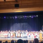 (hrvatski) All the performers of the 50th-anniversary concert in Croatian folk costumes lined up across the stage, with the school banner beside the podium.