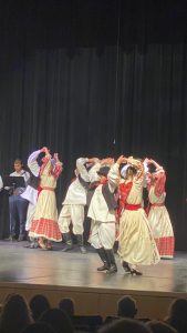 Dancers in white-and-red costumes with arms raised in a turn, a tamburaš at a music stand behind them.