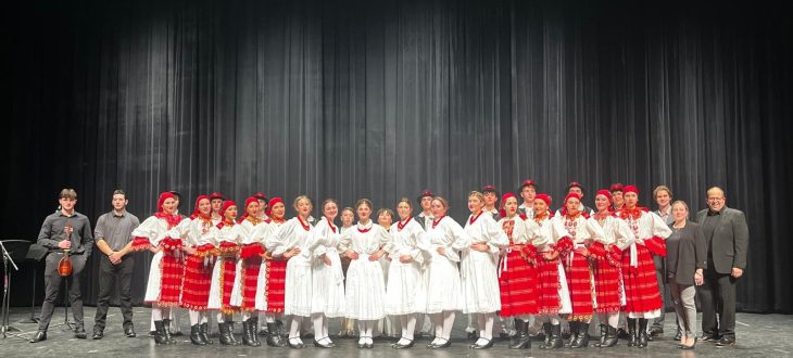Dancers in red-and-white Posavina-style folk costumes with red headscarves and hats line up across a theater stage after the 2025 Annual Concert, with tamburaši and adults standing at the sides.