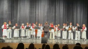 A tamburica orchestra in Croatian folk costumes and vests plays behind music stands on a theater stage, led by a conductor, with the audience in the foreground.