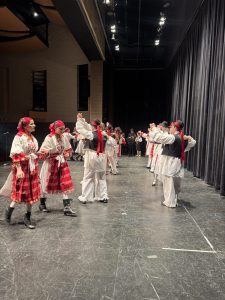 Couples in red-and-white folk costumes, the men in black vests and hats with red ribbons, dance arm in arm across the stage.