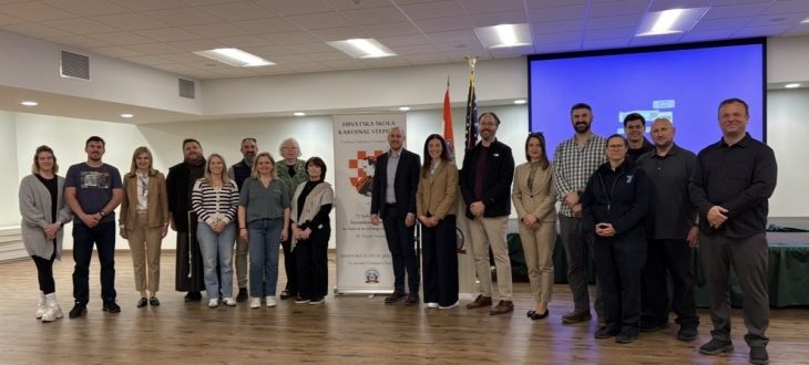 About twenty adults stand together in a parish hall beside the Hrvatska škola Kardinal Stepinac roll-up banner, with the Croatian and American flags behind them.