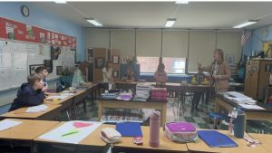 A workshop leader talks with older students seated around desks in a bright classroom.