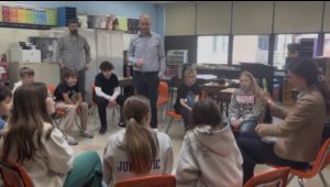 Older students sit in a circle of chairs during a workshop while a workshop leader speaks and two adults look on.