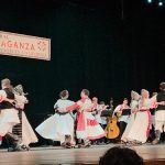 Dancers in Croatian folk costumes and beaded headpieces dancing a kolo on stage beneath the "Croatian Cultural Extravaganza, Los Angeles, California" banner, tamburaši playing behind them.