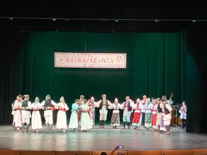 (English) A wide view of the whole group dancing a kolo across the stage under the Extravaganza banner.