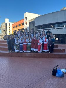 (English) Klub Stepinac performers in nošnja with parents and chaperones, posed on the steps of the plaza outside the venue on a sunny day.