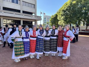 (English) Girls in colorful Croatian folk costumes and beaded headpieces smiling together on the plaza before the concert.