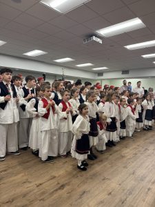 A large group of students in a variety of Croatian folk costumes stands in the parish hall, with parents watching from the back.