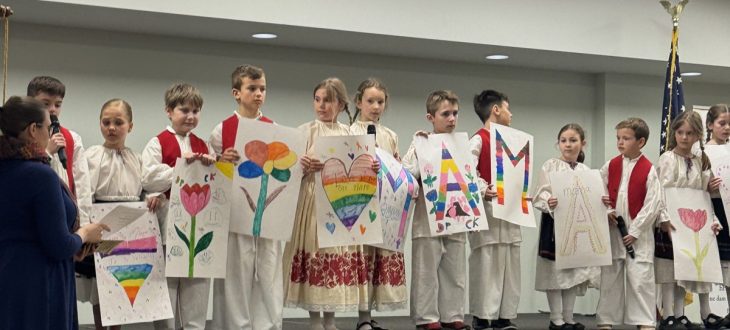 The youngest students in Croatian folk costumes stand on a small stage holding hand-drawn posters of hearts, flowers and rainbows, with letters spelling MAMA, while a teacher stands beside the stage with the program in hand.