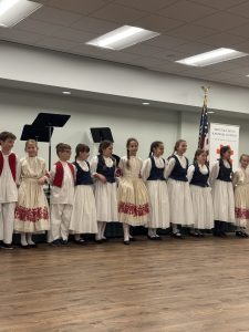 A row of girls and boys in Croatian folk costumes, the girls in white dresses with blue vests or red-embroidered skirts, performs in front of the school banner.