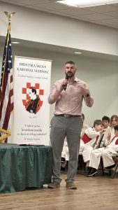 A man speaks into a microphone beside the Hrvatska škola Kardinal Stepinac banner, which shows Blessed Alojzije Stepinac and the quote "U ljubavi prema hrvatskom narodu ne dam se ni od koga natkriliti!", with costumed students seated behind him.